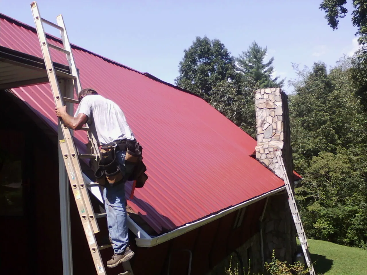 Roofer on a ladder beside a red metal roof and stone chimney — Soffit & Fascia Repair by Roofing Brothers in Royal Oak